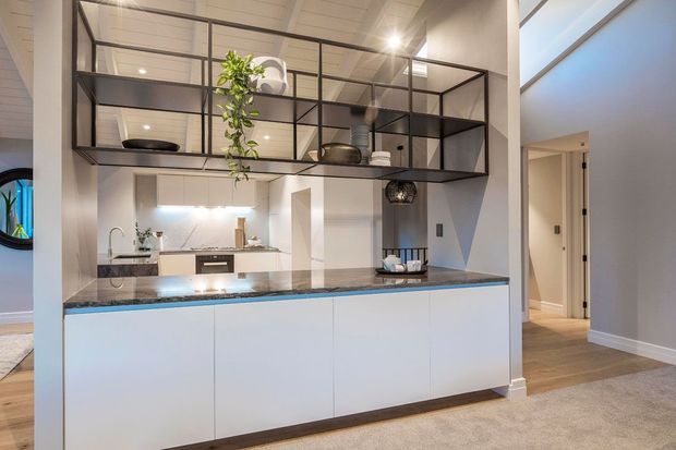 Bright contemporary open-plan kitchen with a large white island, dark stone benchtop, integrated appliances, and black-framed open shelving suspended above the island. The kitchen connects to a carpeted living area and hallway, with high ceilings and clerestory-style glazing.