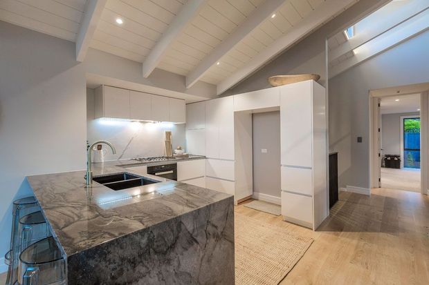 Bright contemporary kitchen with a large marble-look island and integrated sink, white flat-panel cabinetry, stainless appliances, and subtle under-cabinet lighting. The room features a painted white timber-lined vaulted ceiling with exposed beams, light timber flooring, and a hallway leading to adjacent spaces.