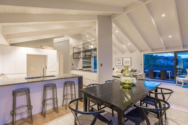 Open-plan contemporary interior combining a kitchen, dining area and living space. The room features a large dark dining table with black chairs, a white kitchen with island and bar stools, exposed white-painted ceiling beams, a high sloped ceiling, and extensive glazing opening to an outdoor deck.