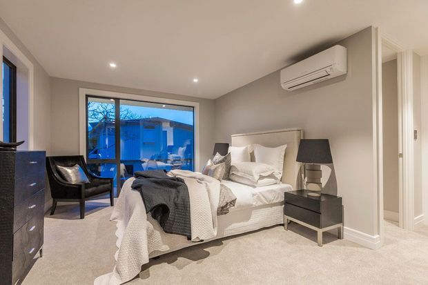 A contemporary bedroom with a neutral palette, upholstered bed, layered white bedding, dark bedside tables and lamps, carpeted flooring, recessed ceiling lights, and a large glazed opening overlooking the exterior. A wall-mounted split-system air conditioner is visible, with a hallway or adjoining space beyond the open doorway.