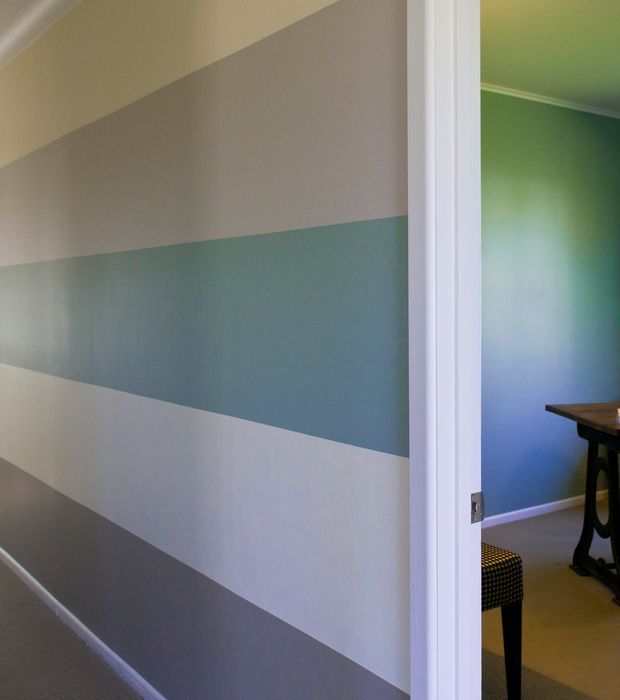 Interior view of a hallway with broad horizontal painted bands in muted grey, blue-green, and off-white. A white-trimmed doorway opens to a blue-green room containing part of a dark table and upholstered seating. The space has simple painted walls, carpeted flooring, and traditional white trim.