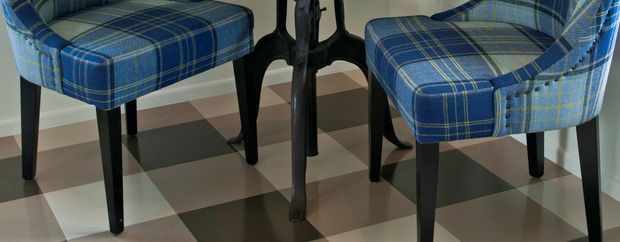 Close-up of a small dining area showing blue plaid upholstered dining chairs with dark wooden legs around a dark table, set on a large black, white, and grey checkerboard floor. The image focuses on furniture, upholstery, and flooring details.