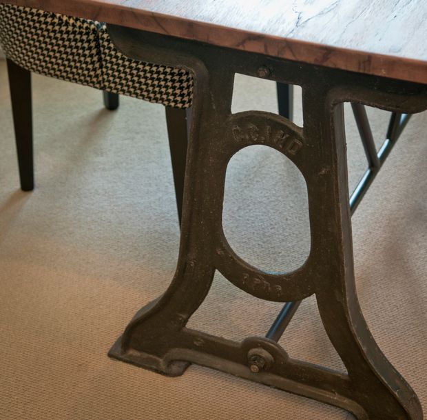 Close-up detail of an antique cast-iron table base with arched cut-outs and curved supports, paired with a dark wood tabletop and a black-and-cream patterned upholstered chair. The setting appears to be an interior dining or living area with neutral carpet and subdued, warm tones.