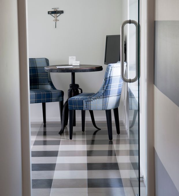 A compact, refined dining or café-style seating area viewed through a glass door. Two blue plaid upholstered chairs surround a small round dark timber table with a central pedestal base. The room has a bold black, white and grey checkerboard floor, pale walls, a dark wall-mounted screen or artwork, and a glass entry door with a prominent metal pull handle. The space combines classic furniture with a crisp contemporary setting.