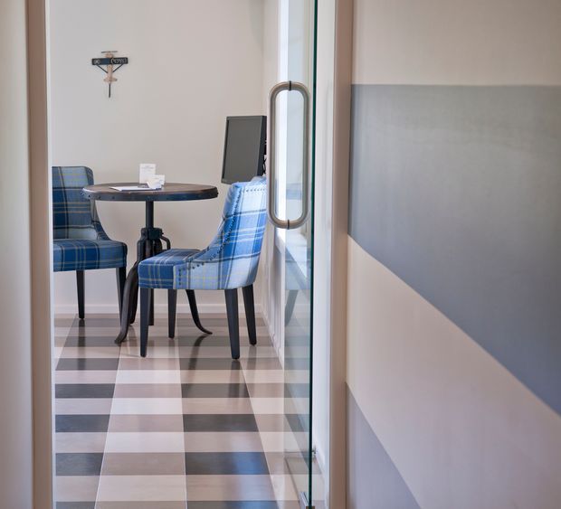 A compact, refined dining nook viewed through a glazed doorway, furnished with a small round dark table and blue plaid upholstered chairs. The room features a bold large-scale checkerboard floor in pale grey, white, and dark grey, light walls with grey geometric wall treatment, and a glass door with a tall metal pull handle. A small wall-mounted decorative object and a dark screen or artwork are visible in the background.