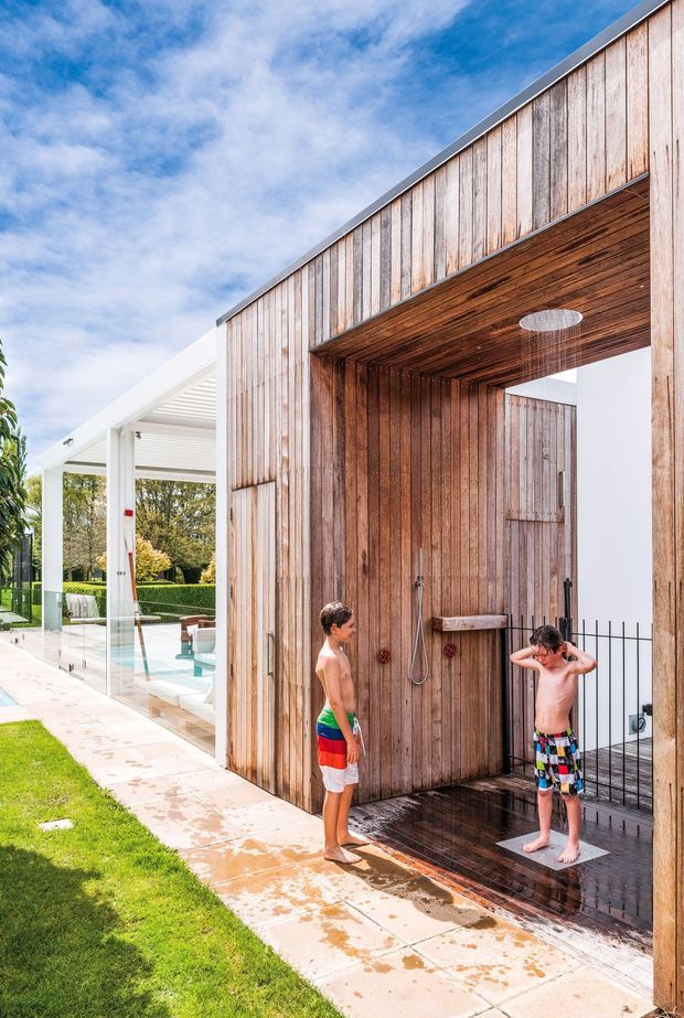 Exterior view of a contemporary poolside outdoor shower enclosure clad in vertical weathered timber, with a timber-lined recessed shower bay and built-in shower fittings. A white-framed glazed pool pavilion and swimming pool are visible beside a paved path and lawn. Two children are present, but the architectural focus is the outdoor shower and pool setting.
