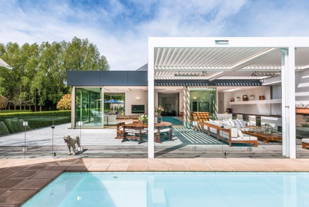Contemporary poolside entertaining area with a white-framed glazed pavilion, open louvred pergola roof, outdoor lounge seating, dining area, and integrated bar. The glass-walled structure opens directly onto a tiled swimming pool terrace and landscaped garden, with strong geometric shadows and abundant daylight.