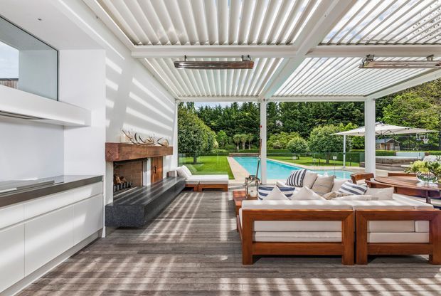 A bright, open-plan covered outdoor living area with a white louvred pergola roof, timber decking, built-in cabinetry and a fireplace along the left wall, and substantial timber-framed outdoor lounge seating. The space opens directly onto a landscaped garden and turquoise swimming pool, with strong striped sunlight and shadows cast through the louvres.