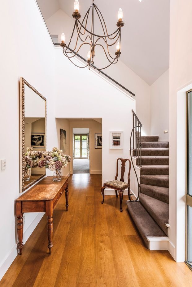 A bright, elegant residential entry hall with polished timber flooring, white walls, a tall vaulted ceiling, and a carpeted staircase with a dark metal decorative balustrade. A traditional timber console table, ornate framed mirror, antique chair, artwork, and a large sculptural chandelier create a refined classic character. A doorway leads toward a glazed exterior entrance at the rear.