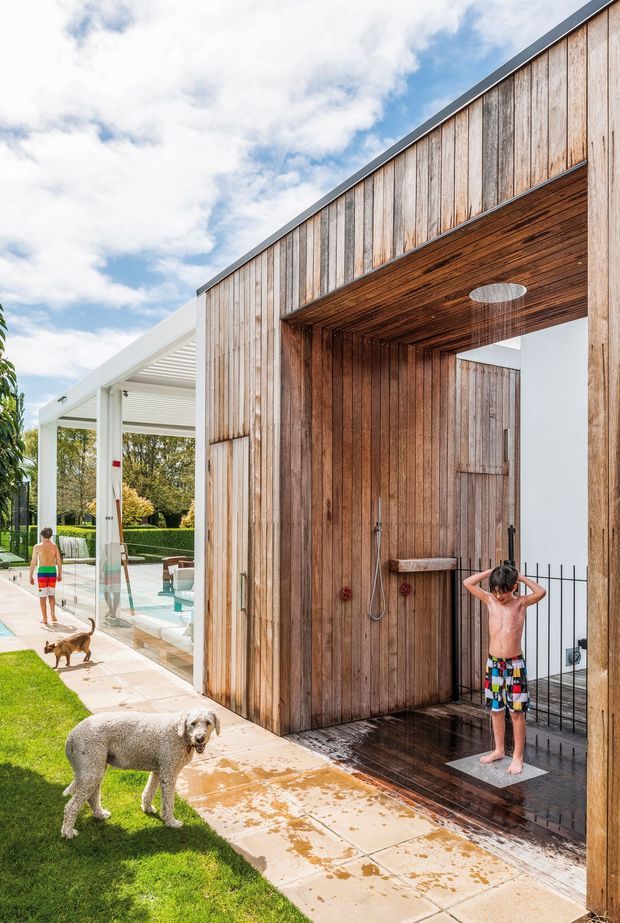 Exterior view of a contemporary poolside outdoor shower enclosure clad in vertical weathered timber boards. The shower has a timber-lined ceiling and walls, exposed plumbing fixtures, a circular ceiling showerhead, and dark tiled flooring. A white covered patio or pool structure, paved walkway, lawn, and garden are visible alongside it.