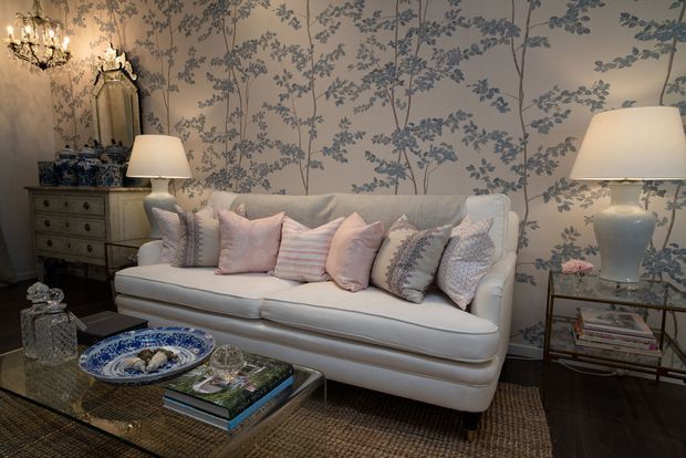 Elegant, traditionally styled living room featuring a cream upholstered sofa with layered pastel cushions, botanical blue-and-white wallpaper, antique-style chest and mirror, crystal chandelier, table lamps, glass side table, and a woven area rug over dark timber flooring.
