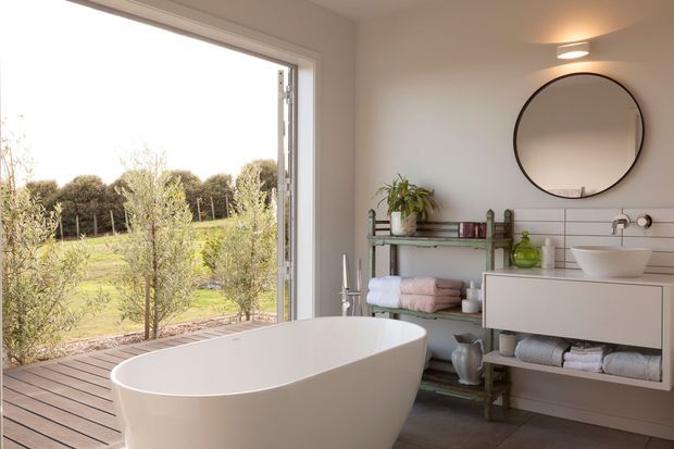 Bright contemporary bathroom with a freestanding white bathtub positioned beside large floor-to-ceiling glazing, overlooking a landscaped rural garden. The room includes a wall-mounted double vanity with a white vessel basin, round mirror, open timber shelving with folded towels and toiletries, recessed ceiling lighting, and dark tiled flooring. A timber deck extends outside the glazing.