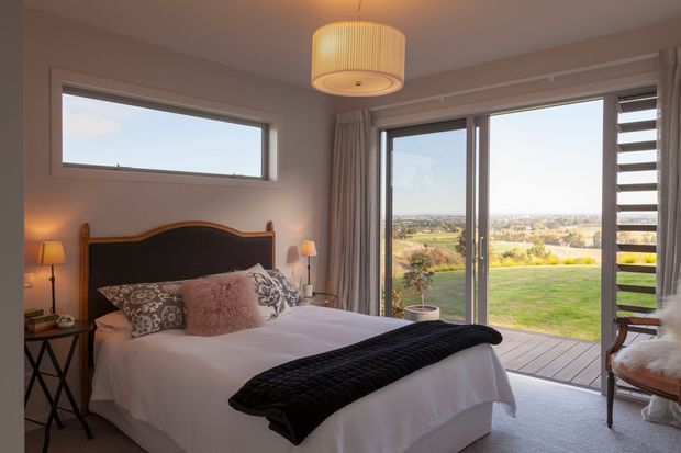 A warmly lit bedroom with a queen or king bed, upholstered dark headboard, layered patterned and plush cushions, bedside tables and lamps, and a black textured throw. Full-height glazed doors open to a timber deck and expansive landscaped rural or suburban views. A high horizontal window and louvred glazing provide additional daylight and ventilation. The room has a calm, refined transitional feel.