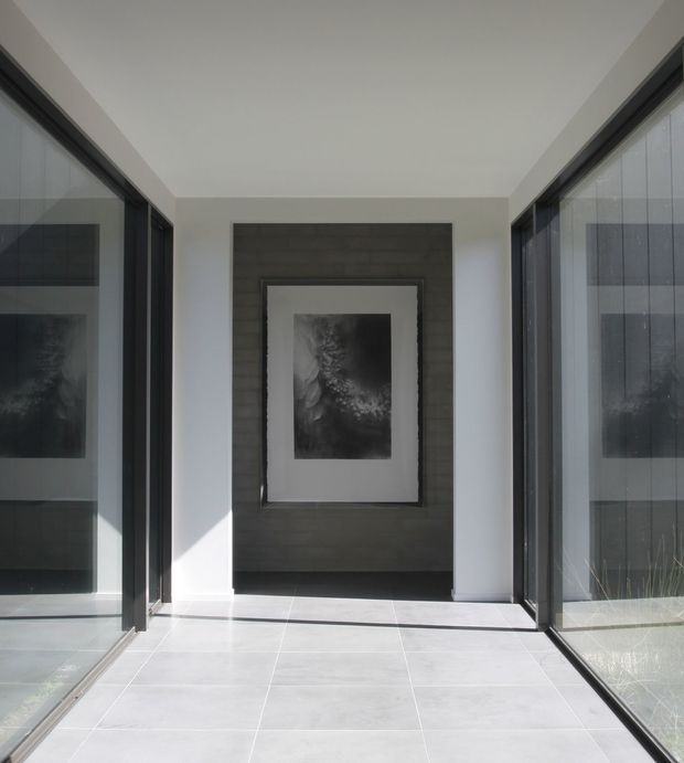 A bright, minimalist glazed hallway or gallery passage with full-height dark-framed glass walls on both sides, pale large-format tiled flooring, white walls and ceiling, and a dark textured feature wall displaying a monochrome artwork at the far end. Strong natural light creates sharp shadows across the floor.