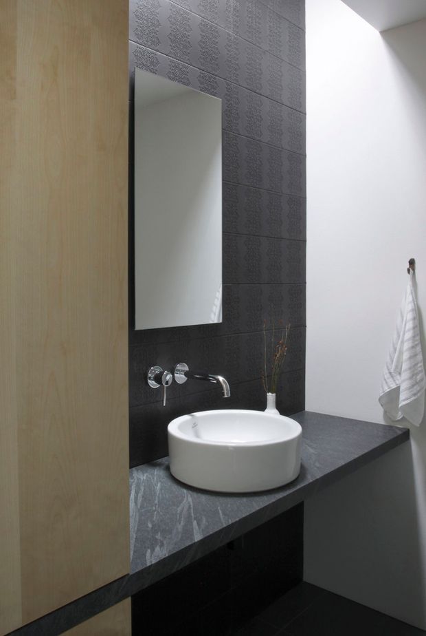 Compact contemporary bathroom vanity with a white vessel basin, wall-mounted chrome tapware, large frameless mirror, dark patterned tile feature wall, and dark stone benchtop. Light timber cabinetry partially frames the left side, while a white towel hangs on the right wall.