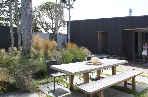 A contemporary outdoor dining area beside a dark vertically clad, low-profile building. A long timber dining table with matching benches and black chairs sits on large concrete pavers, surrounded by ornamental grasses and mature trees. An open doorway connects to the building interior, while the landscaping creates a natural, private setting.