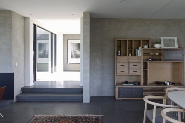 Contemporary interior featuring a dark tiled floor, raw grey concrete-look walls and columns, and extensive custom timber built-in shelving and cabinetry. A raised hallway with framed artwork leads toward glazed doors, while a dining table and curved chairs occupy the foreground.