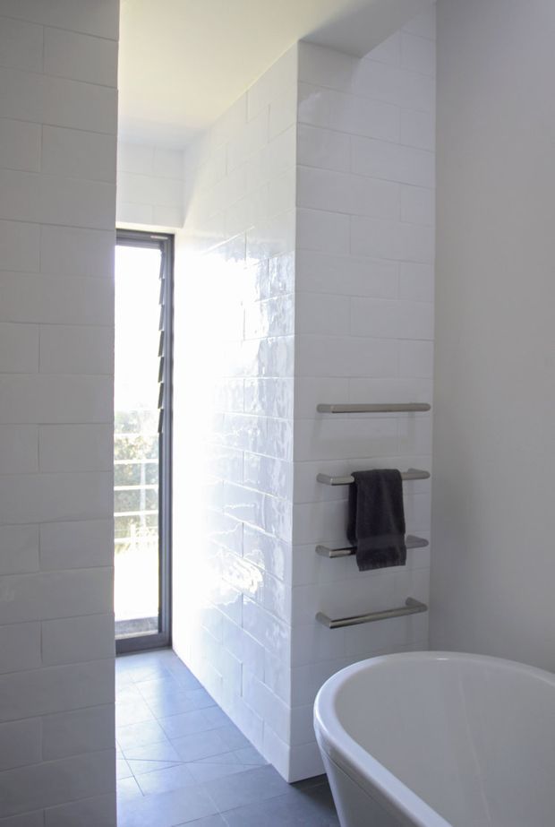 A bright, minimalist bathroom with white rectangular wall tiles, pale floor tiles, a freestanding white bathtub, a narrow vertical window, and a wall-mounted brushed metal towel rail holding a dark towel. The room has clean geometric lines and abundant natural light.
