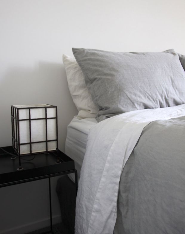 Close-up of a simply styled bedroom corner with white bedding, layered light-grey pillows and a dark bedside table. A small rectangular grid-frame bedside lamp sits on the table against a plain light wall, creating a restrained monochromatic composition.