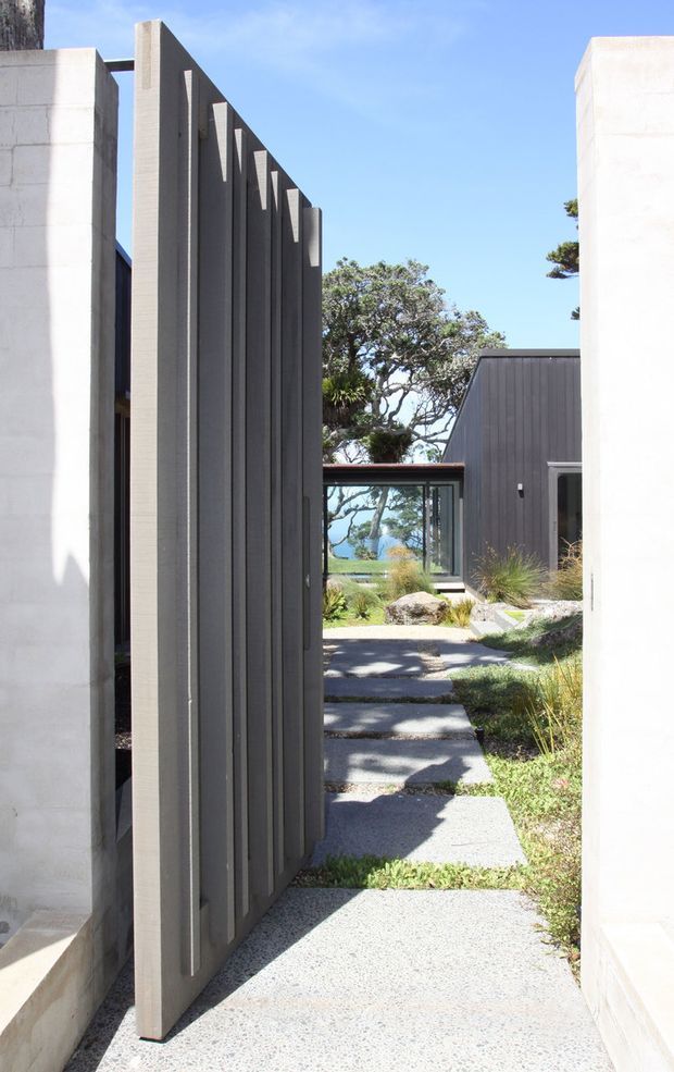 Exterior architectural view through a narrow entry passage, featuring a tall pivoting or hinged dark grey vertical-slat screen door set between smooth white rendered masonry walls. A stepping-stone path leads through a landscaped courtyard toward a dark-clad pavilion with glazing, with coastal vegetation and blue water visible beyond. The composition is minimalist and strongly geometric, with sharp sunlight and deep shadows.