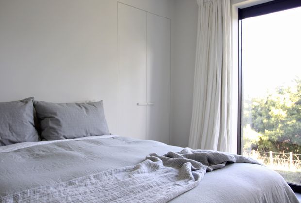 A bright, minimalist bedroom with a neatly made bed dressed in layered light-grey linen, integrated flush wall storage, pale curtains, and a large dark-framed window overlooking greenery.