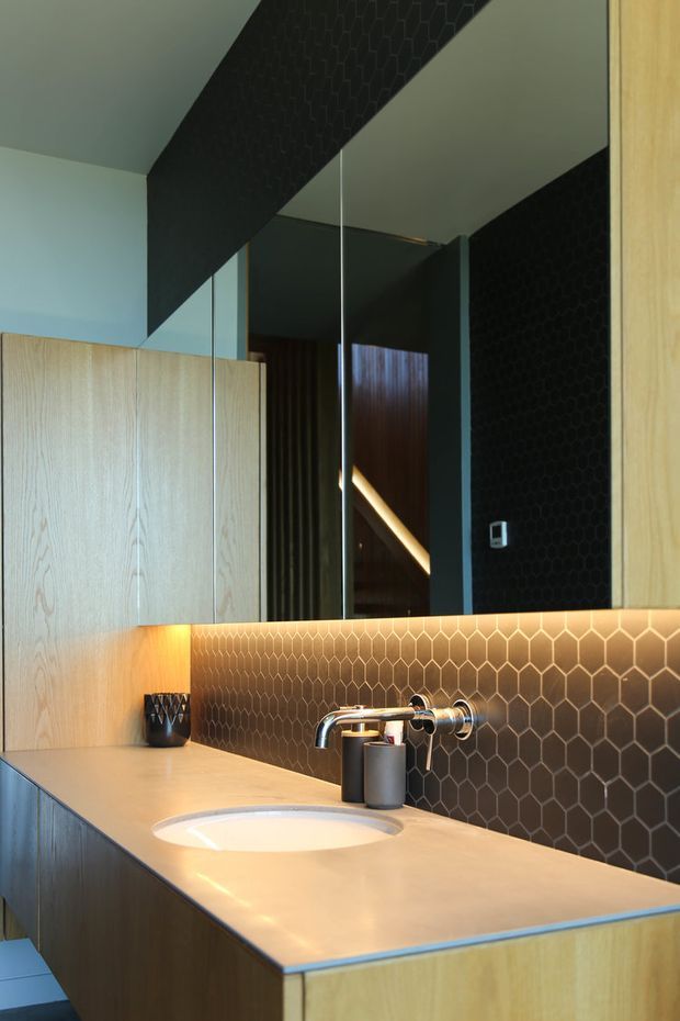 Close-up view of a contemporary bathroom vanity with a pale stone benchtop, integrated white basin, wall-mounted chrome tapware, timber-grain cabinetry, and a dark brown hexagonal tile splashback. A large mirror reflects the room, while warm under-cabinet lighting highlights the textured tile surface.