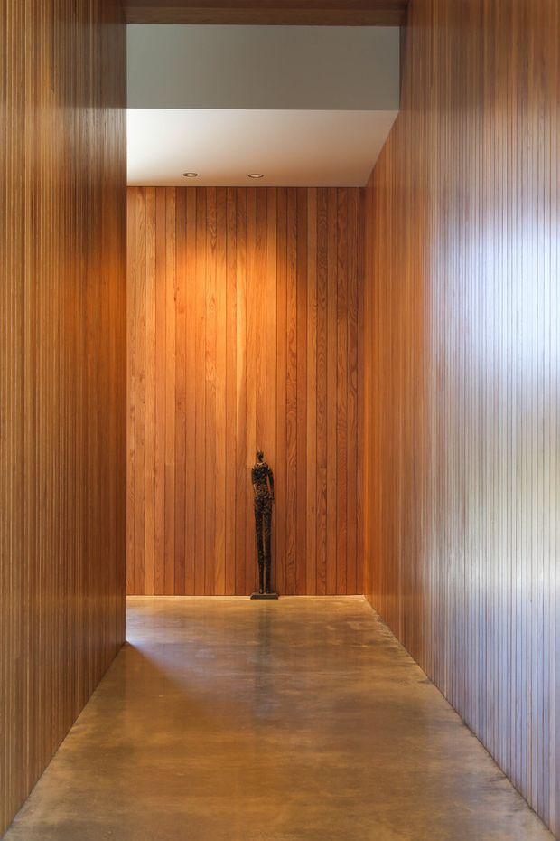 A narrow, tall interior passage or entry hall lined on both side walls and the end wall with vertical warm-toned timber panelling. The space has a smooth grey-brown concrete floor, a simple white recessed ceiling with downlights, and a small dark sculptural figure positioned at the far end. The composition is highly symmetrical and minimalist, with strong linear perspective and warm ambient lighting.