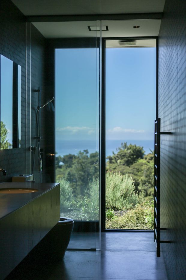 A dark, contemporary bathroom with a walk-in glass shower, wall-mounted shower fittings, integrated vanity, and toilet. Small dark textured wall tiles contrast with a smooth concrete-look floor and vanity surfaces. Full-height glazing or a glass door opens toward a landscaped garden with a distant ocean view, bringing strong natural light into the otherwise moody interior.
