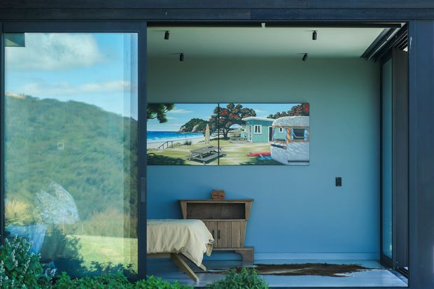 A compact blue bedroom viewed through large dark-framed sliding glazing, with a low bed, built-in or freestanding timber storage unit, recessed track lighting, and a large beach-themed artwork. The room opens visually to a lush, mountainous landscape through the glazing, creating a strong indoor-outdoor connection. The interior has a clean, contemporary feel with bold blue walls and dark framing.