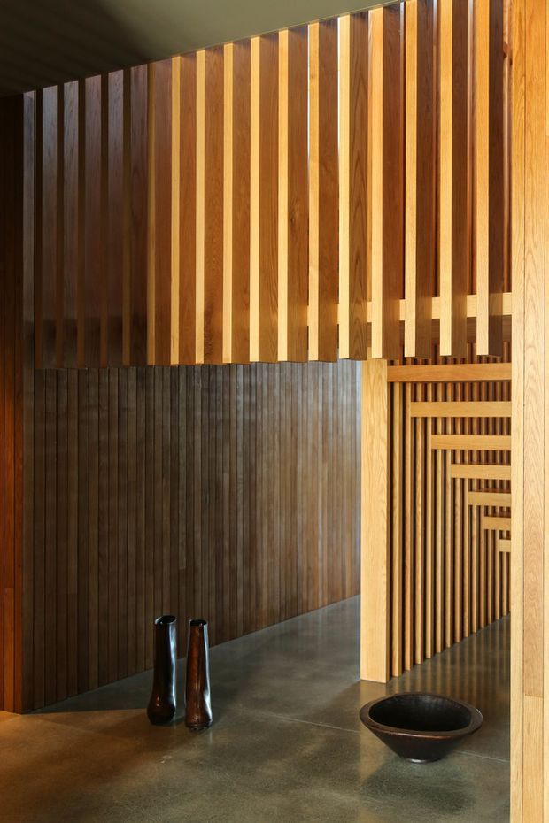 A warm, highly crafted interior featuring extensive vertical timber battens and slatted screens forming layered openings and a corridor-like passage. Dark timber wall panelling contrasts with lighter golden timber fins and a smooth polished concrete floor. Minimal sculptural black vessels sit on the floor, while a dark ceiling and deep shadows create a quiet, gallery-like atmosphere.