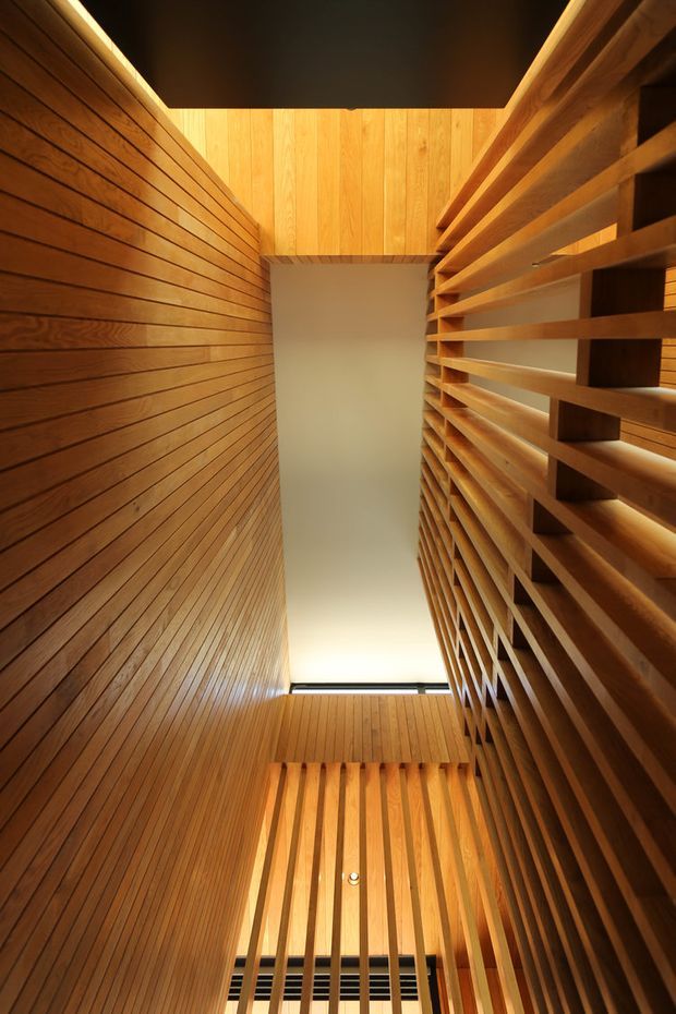 Vertical interior view of a tall, narrow timber-lined architectural space, likely a stairwell or circulation void. Warm wood slat wall panelling covers the left and lower walls, while the right side features a substantial open timber screen or balustrade with horizontal slats. A high ceiling opening or skylight brings in soft light, emphasizing the verticality and layered wood construction. The space has a strongly crafted, contemporary architectural character.