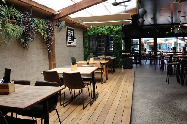 A contemporary restaurant or café interior with timber-framed glazing, exposed timber beams, a planted feature wall, concrete block walls, timber dining tables, dark chairs, pendant lighting, and a polished concrete floor. The space includes a raised timber dining platform and opens toward an outdoor seating area.