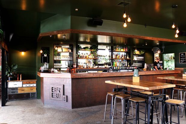 A dark, moody bar interior with a long timber-fronted counter, polished concrete floor, open shelving stocked with bottles and glassware, pendant lighting, bar stools, and a high communal table. Green-painted walls and ceiling contrast with warm timber detailing and industrial-style metal furniture.