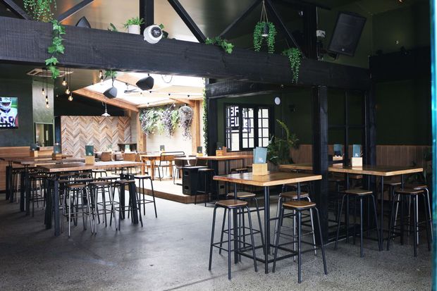 A spacious contemporary hospitality interior with a dark, industrial-inspired fit-out. The venue features high communal tables with black metal stools, exposed dark structural beams, hanging greenery, polished concrete flooring, timber dining tables, black-framed glazing, and a bright rear dining area with a chevron timber feature wall and abundant planting.