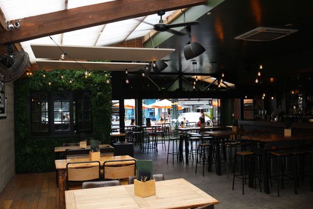 Interior view of a dark, industrial-style restaurant or bar with timber dining tables, black metal stools and chairs, a long bar counter, pendant and track lighting, exposed timber beams, ceiling fans, and a green living-wall feature around a window. Large glazed openings connect visually to an outdoor dining area.