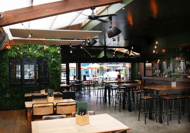 A spacious industrial-style bar and dining venue with dark ceilings, exposed timber beams, hanging pendant lights, ceiling fans, and a mix of high bar tables and lower timber dining tables. A greenery-covered feature wall and black-framed window add visual interest, while a long timber bar with displayed bottles occupies the right side. The open frontage leads toward an outdoor street seating area.