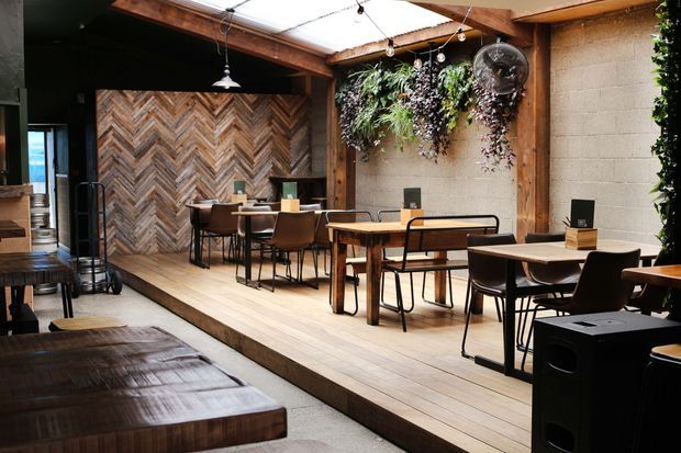 A warm, industrial-style commercial dining space with timber decking, wooden tables and chairs, a chevron-patterned timber feature wall, exposed timber beams, hanging pendant lights, lush vertical planting, and a translucent roof creating a bright indoor-outdoor atmosphere.