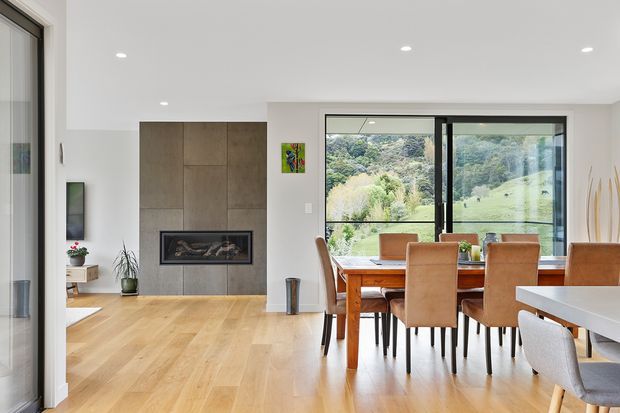 Bright contemporary open-plan dining and living area with warm timber flooring, a large timber dining table and upholstered dining chairs positioned beside expansive black-framed glazing. A minimalist concrete-toned feature wall incorporates a built-in fireplace, with landscaped greenery visible outside.
