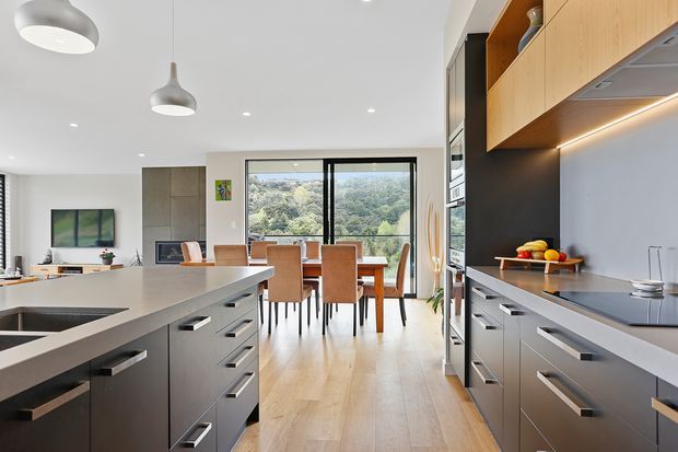 A bright, contemporary open-plan kitchen with a large island, dark lower cabinetry, warm timber overhead cabinetry, integrated appliances, and a dark splashback. The kitchen opens to a dining and living area with expansive glazing overlooking leafy greenery. Light timber flooring, streamlined pendant lights, and recessed ceiling lights create a clean, refined feel.