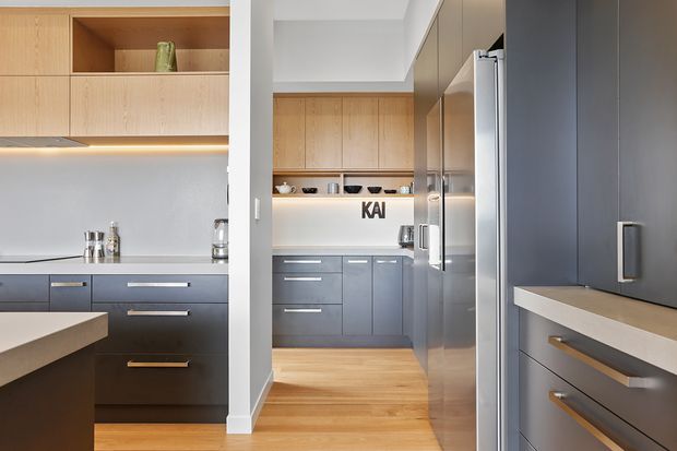 Contemporary kitchen featuring warm timber upper cabinetry, matte dark grey lower cabinetry, integrated appliances, pale benchtops, and light timber flooring. The space is viewed through a wide opening, with layered cabinetry and concealed under-cabinet lighting creating a clean, refined look.