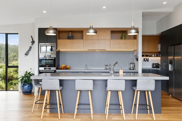 A bright contemporary kitchen with a large island and four upholstered bar stools. The space features flat-panel black cabinetry, warm timber overhead joinery, a grey island and splashback, integrated stainless-steel appliances, pendant lights, and timber flooring. Floor-to-ceiling glazing opens to a leafy view at the side.