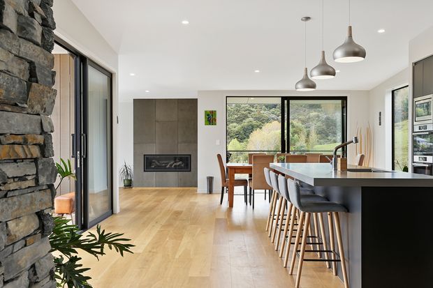 A bright open-plan contemporary interior combining a kitchen, dining area and living space. The room features a long dark kitchen island with timber bar stools, a timber dining table, pendant lights, pale timber flooring, large black-framed windows and doors, and a substantial exposed-stone fireplace wall with a built-in firebox.