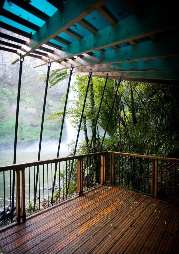 A covered elevated terrace or deck overlooking a lush forested gorge and waterfall. The deck has a warm timber-toned grid floor, timber handrails with dark metal balusters, and a translucent roof supported by exposed timber beams and slender angled structural posts. Dense native vegetation surrounds the open sides, creating a strongly integrated nature setting.