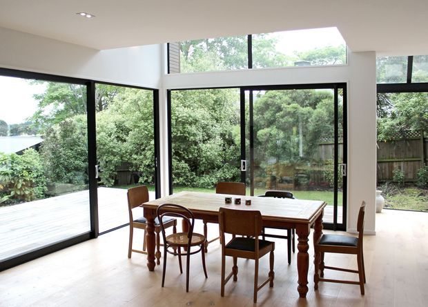 Bright open-plan dining area with a traditional timber dining table and mixed wooden chairs, set beside extensive black-framed glazing. The room opens to a timber deck and leafy garden, with high clerestory windows bringing in abundant natural light. Pale flooring and walls create an airy, warm interior.