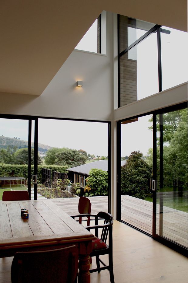 A contemporary open-plan dining area with a large timber dining table and chairs, framed by extensive floor-to-ceiling black-framed glazing. The room opens onto a timber deck and landscaped garden, with views toward a green, hilly setting. A dramatic angular ceiling and double-height glazed corner create a spacious, light-filled interior.