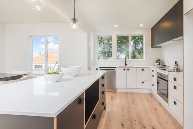 Bright contemporary kitchen with white cabinetry, a large white island, pale timber flooring, black hardware and dark upper cabinets. Multiple windows provide abundant natural light and views to neighbouring homes and trees, while pendant and recessed lighting illuminate the space.