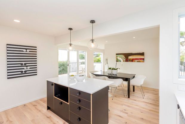 Bright open-plan kitchen and dining area with a central dark-wood island featuring a glossy white benchtop, integrated drawers and shelving. Pale timber flooring, white walls and ceiling, pendant lights, contemporary artwork, and a dining table with upholstered chairs create a clean, airy interior. Large windows and glazed doors provide natural light and views to greenery outside.