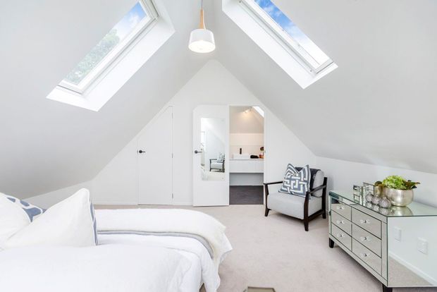 Bright attic-style bedroom with a steeply sloped white ceiling, two skylights, white bedding, pale carpet, and a grey upholstered accent chair. A mirrored drawer chest and small decorative accessories sit along the right wall, while an open door reveals a compact bathroom or dressing area beyond.