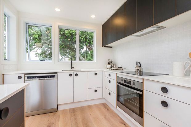 Bright contemporary kitchen with white flat-panel cabinetry, dark overhead cupboards, white tiled splashback, stainless-steel dishwasher and oven, pale timber flooring, and large windows overlooking greenery. A compact peninsula or island is visible at left.