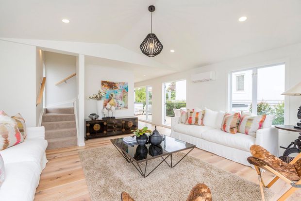 Bright, airy contemporary living room with white sofas, colourful patterned cushions, a black glass coffee table, textured rug, pale timber flooring, and large windows and glazed doors. An open stairway and landing are visible at the rear, along with a dark geometric pendant light and decorative sideboard.